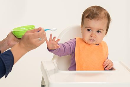 A baby in a high chair looks unhappily at some food that is being offered by a nearby adult holding a bowl and spoon.