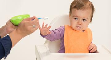 A baby in a high chair looks unhappily at some food that is being offered by a nearby adult holding a bowl and spoon.