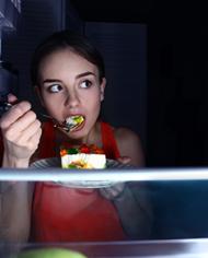 A woman standing in front of an open refrigerator in the dark eating.